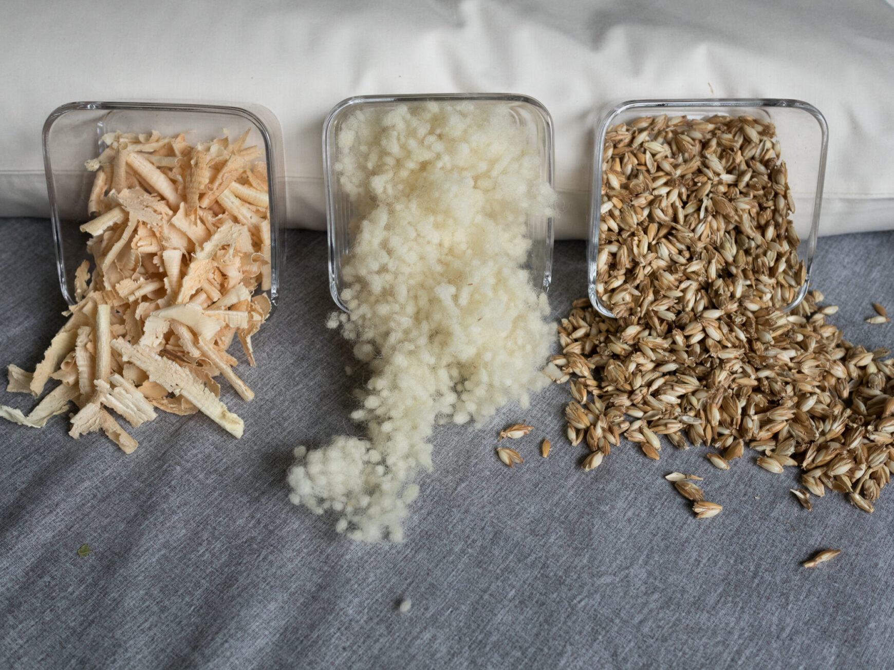 Dinkelspelzen, Schafswolle und Zirbenspäne in Glasschüsseln an ein Körnerkopfkissen angelehnt. Dinkelfüllung, Schafswollefüllung und Zirbenfüllung für Körnerkopfkissen.